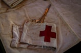 A pair of scissors lies on a fabric surface, partially covered by a piece of cloth featuring a red cross symbol. The cloth appears slightly worn and stained, suggesting frequent use, possibly in a medical or emergency setting. The scene is dimly lit, casting shadows and creating a somewhat somber atmosphere.