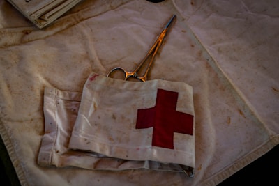A pair of scissors lies on a fabric surface, partially covered by a piece of cloth featuring a red cross symbol. The cloth appears slightly worn and stained, suggesting frequent use, possibly in a medical or emergency setting. The scene is dimly lit, casting shadows and creating a somewhat somber atmosphere.