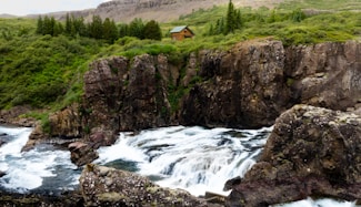 A rustic cabin is nestled among lush, green trees atop a rocky cliff. Below, a river cascades over rocks, creating frothy white rapids. In the background, hills stretch into the distance, covered with vegetation.