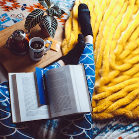 A cozy setup with a teapot, cup of herbal tea, and an open book on a cream-colored blanket.