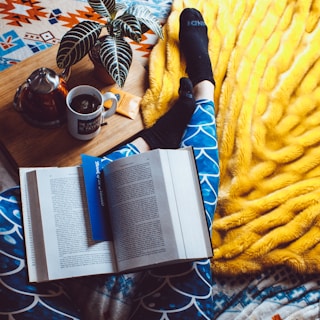 A cozy setting featuring an open book with a blue bookmark resting on patterned blue leggings. A person is wearing black socks, lounging on a bright yellow blanket. Nearby, a wooden tray holds a potted plant, a cup of tea, a teapot, and a small snack.