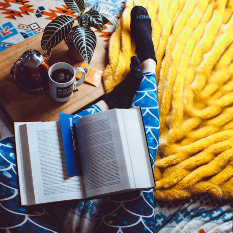 A cozy reading nook with a blue blanket and a wellness book beside a steaming cup of tea.
