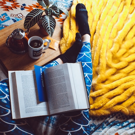 A cozy setting featuring an open book with a blue bookmark resting on patterned blue leggings. A person is wearing black socks, lounging on a bright yellow blanket. Nearby, a wooden tray holds a potted plant, a cup of tea, a teapot, and a small snack.