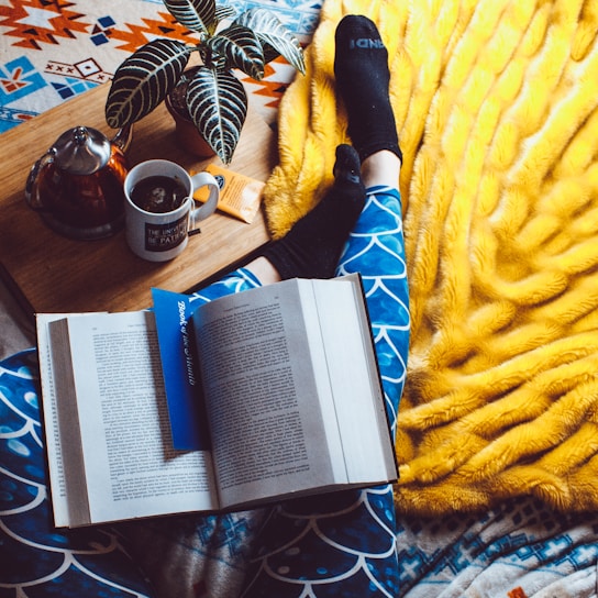 A cozy setting featuring an open book with a blue bookmark resting on patterned blue leggings. A person is wearing black socks, lounging on a bright yellow blanket. Nearby, a wooden tray holds a potted plant, a cup of tea, a teapot, and a small snack.