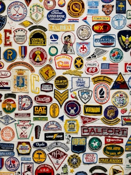 A collage of various colorful embroidered patches displayed against a white background. The patches include different insignias, logos, numbers, and symbols representing organizations, sports teams, clubs, and brands. Some patches have text in bold, while others feature graphic designs and emblems such as flags, stars, animals, and badges.