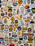 A collage of various colorful embroidered patches displayed against a white background. The patches include different insignias, logos, numbers, and symbols representing organizations, sports teams, clubs, and brands. Some patches have text in bold, while others feature graphic designs and emblems such as flags, stars, animals, and badges.