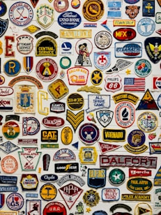 A collage of various colorful embroidered patches displayed against a white background. The patches include different insignias, logos, numbers, and symbols representing organizations, sports teams, clubs, and brands. Some patches have text in bold, while others feature graphic designs and emblems such as flags, stars, animals, and badges.
