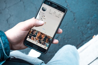 A person holding a smartphone displaying the Instagram profile page. The screen shows personal information and photo thumbnails. The background includes a textured, dark pavement and part of the person's denim jacket and light-colored pants.