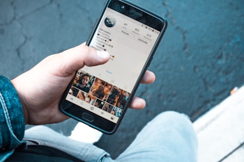 A person holding a smartphone displaying the Instagram profile page. The screen shows personal information and photo thumbnails. The background includes a textured, dark pavement and part of the person's denim jacket and light-colored pants.