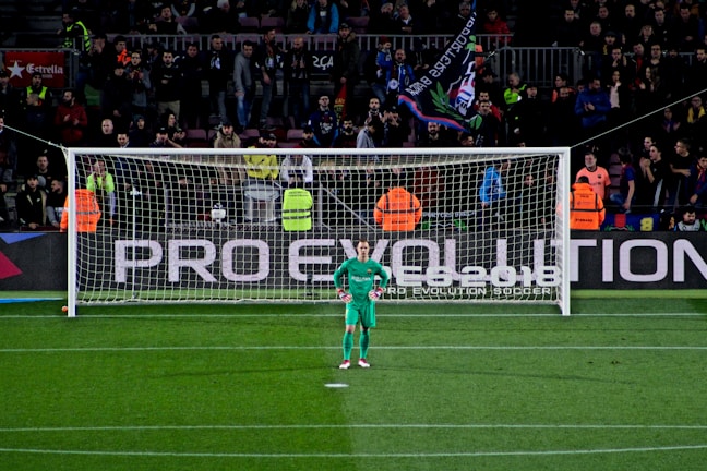 A football goalkeeper stands in front of a goalpost on a soccer field. The audience is filled with spectators, some wearing brightly colored clothing, while security personnel in fluorescent vests stand nearby. A large banner with the words 'PRO EVOLUTION SOCCER' is visible behind the goal.