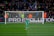 A football goalkeeper stands in front of a goalpost on a soccer field. The audience is filled with spectators, some wearing brightly colored clothing, while security personnel in fluorescent vests stand nearby. A large banner with the words 'PRO EVOLUTION SOCCER' is visible behind the goal.