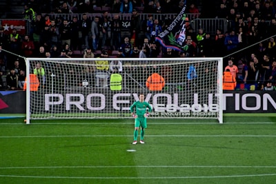 A football goalkeeper stands in front of a goalpost on a soccer field. The audience is filled with spectators, some wearing brightly colored clothing, while security personnel in fluorescent vests stand nearby. A large banner with the words 'PRO EVOLUTION SOCCER' is visible behind the goal.