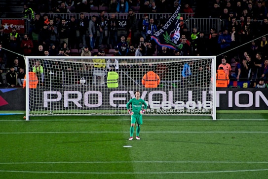 A football goalkeeper stands in front of a goalpost on a soccer field. The audience is filled with spectators, some wearing brightly colored clothing, while security personnel in fluorescent vests stand nearby. A large banner with the words 'PRO EVOLUTION SOCCER' is visible behind the goal.