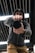 A person wearing a hoodie and cap is holding a camera up close to the lens, pointing directly at the viewer. The perspective is dynamic and low-angle, capturing a sense of immediacy. The background features industrial ceiling elements with linear patterns and bright lighting.