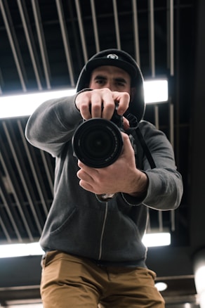 A person wearing a hoodie and cap is holding a camera up close to the lens, pointing directly at the viewer. The perspective is dynamic and low-angle, capturing a sense of immediacy. The background features industrial ceiling elements with linear patterns and bright lighting.