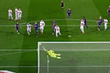 A soccer match with players from two teams, one in blue and red uniforms and the other in white with red stripes. A goalkeeper in a bright yellow uniform is diving to save a penalty kick while other players watch from around the penalty area.