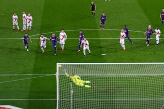 A soccer match with players from two teams, one in blue and red uniforms and the other in white with red stripes. A goalkeeper in a bright yellow uniform is diving to save a penalty kick while other players watch from around the penalty area.