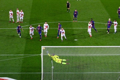 A soccer match with players from two teams, one in blue and red uniforms and the other in white with red stripes. A goalkeeper in a bright yellow uniform is diving to save a penalty kick while other players watch from around the penalty area.