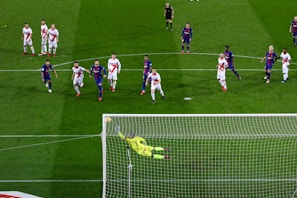 A soccer match with players from two teams, one in blue and red uniforms and the other in white with red stripes. A goalkeeper in a bright yellow uniform is diving to save a penalty kick while other players watch from around the penalty area.