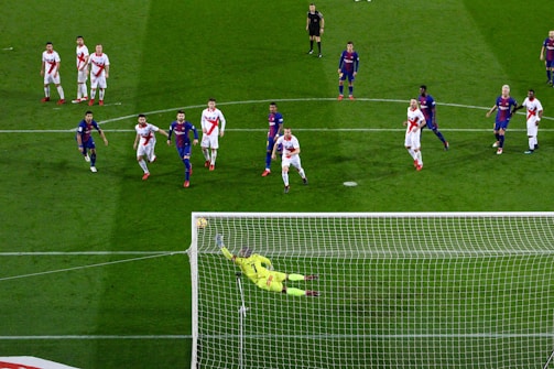 A soccer match with players from two teams, one in blue and red uniforms and the other in white with red stripes. A goalkeeper in a bright yellow uniform is diving to save a penalty kick while other players watch from around the penalty area.