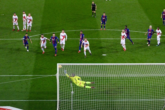 A soccer match with players from two teams, one in blue and red uniforms and the other in white with red stripes. A goalkeeper in a bright yellow uniform is diving to save a penalty kick while other players watch from around the penalty area.