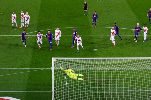 A soccer match with players from two teams, one in blue and red uniforms and the other in white with red stripes. A goalkeeper in a bright yellow uniform is diving to save a penalty kick while other players watch from around the penalty area.
