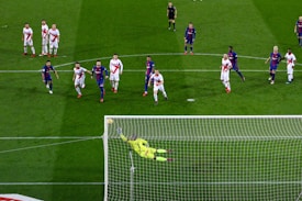 A soccer match with players from two teams, one in blue and red uniforms and the other in white with red stripes. A goalkeeper in a bright yellow uniform is diving to save a penalty kick while other players watch from around the penalty area.