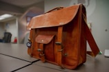 A classic leather briefcase with a rich brown color sits on a modern office desk. The bag features multiple buckles and a small external pocket. The office environment in the background includes a blurred chair and abstract wall art.