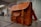 A classic leather briefcase with a rich brown color sits on a modern office desk. The bag features multiple buckles and a small external pocket. The office environment in the background includes a blurred chair and abstract wall art.