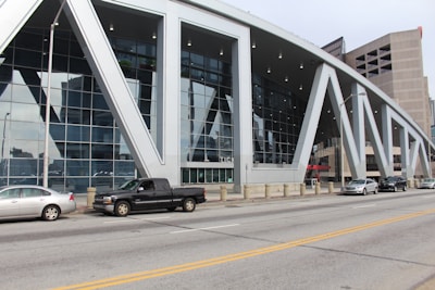 A modern building with a large glass facade and distinctive white geometric supports lining the front. There are several cars parked along the street in front of the building. The structure appears multi-purpose, with tall, sleek windows reflecting nearby structures.
