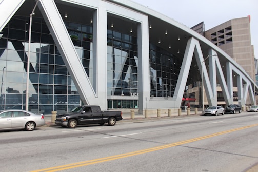 A modern building with a large glass facade and distinctive white geometric supports lining the front. There are several cars parked along the street in front of the building. The structure appears multi-purpose, with tall, sleek windows reflecting nearby structures.