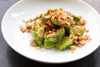 A white plate showcasing a dish of fresh green avocado pieces, topped with a generous amount of crushed nuts and seasoning. The vibrant green contrasts with the light brown of the nuts, providing a visually appealing presentation.