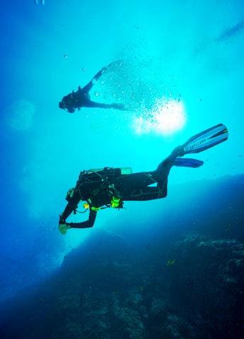 Two scuba divers are submerged in clear blue water, surrounded by bubbles and illuminated by sunlight filtering through the ocean surface. They are exploring an underwater rocky landscape while wearing full diving gear.