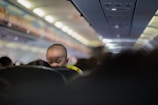 A young child peers over the back of a seat in an airplane cabin, with rows of seats and overhead compartments visible in a dimly lit interior. The child, wearing a yellow outfit, appears curious and slightly amused.