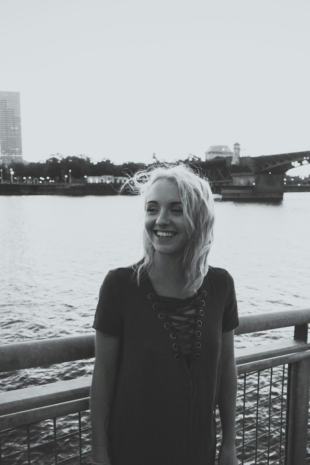 grayscale photo of smiling woman standing near fence above body of water