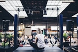 A group of people is gathered in an indoor space, sitting on yoga mats in what appears to be a guided meditation or yoga session. The room is spacious with industrial design elements such as exposed beams and large windows. There is a person seated on a stage facing the group, likely leading the session. Potted plants and sound equipment are present on the stage, creating a serene yet focused atmosphere.