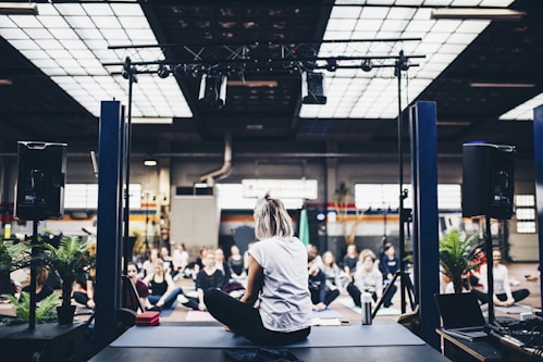 A group of people is gathered in an indoor space, sitting on yoga mats in what appears to be a guided meditation or yoga session. The room is spacious with industrial design elements such as exposed beams and large windows. There is a person seated on a stage facing the group, likely leading the session. Potted plants and sound equipment are present on the stage, creating a serene yet focused atmosphere.