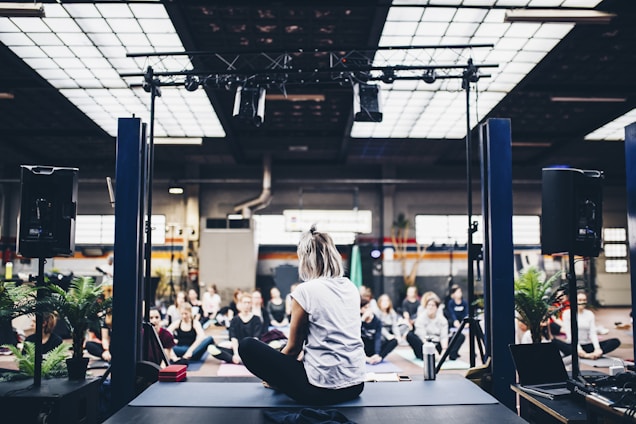 A group of people is gathered in an indoor space, sitting on yoga mats in what appears to be a guided meditation or yoga session. The room is spacious with industrial design elements such as exposed beams and large windows. There is a person seated on a stage facing the group, likely leading the session. Potted plants and sound equipment are present on the stage, creating a serene yet focused atmosphere.