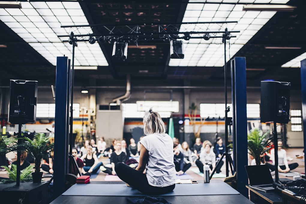 A group of people is gathered in an indoor space, sitting on yoga mats in what appears to be a guided meditation or yoga session. The room is spacious with industrial design elements such as exposed beams and large windows. There is a person seated on a stage facing the group, likely leading the session. Potted plants and sound equipment are present on the stage, creating a serene yet focused atmosphere.