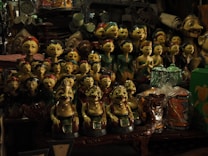 A collection of intricately crafted wooden figurines, each with unique facial expressions and colorful attire, is displayed densely on shelves. Among the figures, there are a variety of handcrafted items and trinkets surrounding them, showcasing a rich assortment of artwork and decor.