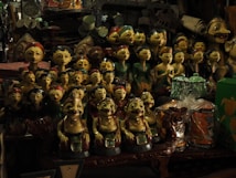 A collection of intricately crafted wooden figurines, each with unique facial expressions and colorful attire, is displayed densely on shelves. Among the figures, there are a variety of handcrafted items and trinkets surrounding them, showcasing a rich assortment of artwork and decor.