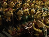 A collection of traditional wooden puppets with intricate facial features, painted in vibrant colors. The puppets are dressed in various traditional costumes, some appearing to be musicians or dancers. The setting suggests a display or market with a focus on cultural craftsmanship.
