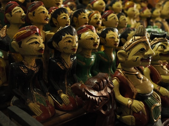 A collection of traditional wooden puppets with intricate facial features, painted in vibrant colors. The puppets are dressed in various traditional costumes, some appearing to be musicians or dancers. The setting suggests a display or market with a focus on cultural craftsmanship.