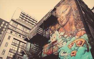 A mural covers the side of a building, featuring a large, intricately detailed face with flowing hair. It is surrounded by abstract elements and vibrant illustrations, including a swimmer and oversized sunglasses. Fire escapes are visible on the side of the building, adding an urban touch.