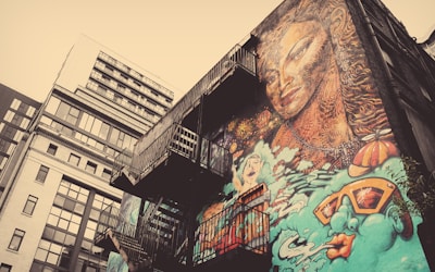 A mural covers the side of a building, featuring a large, intricately detailed face with flowing hair. It is surrounded by abstract elements and vibrant illustrations, including a swimmer and oversized sunglasses. Fire escapes are visible on the side of the building, adding an urban touch.