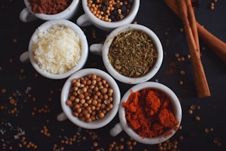Several small white bowls are filled with different spices and ingredients. The bowls hold powdered red spice, coriander seeds, shredded white coconut, dried herbs, and a variety of seeds. Cinnamon sticks are placed beside the bowls, with scattered seeds and spices on a dark surface.
