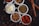 Several small white bowls are filled with different spices and ingredients. The bowls hold powdered red spice, coriander seeds, shredded white coconut, dried herbs, and a variety of seeds. Cinnamon sticks are placed beside the bowls, with scattered seeds and spices on a dark surface.