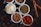 Several small white bowls are filled with different spices and ingredients. The bowls hold powdered red spice, coriander seeds, shredded white coconut, dried herbs, and a variety of seeds. Cinnamon sticks are placed beside the bowls, with scattered seeds and spices on a dark surface.