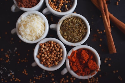 Several small white bowls are filled with different spices and ingredients. The bowls hold powdered red spice, coriander seeds, shredded white coconut, dried herbs, and a variety of seeds. Cinnamon sticks are placed beside the bowls, with scattered seeds and spices on a dark surface.