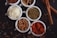 Several small white bowls are filled with different spices and ingredients. The bowls hold powdered red spice, coriander seeds, shredded white coconut, dried herbs, and a variety of seeds. Cinnamon sticks are placed beside the bowls, with scattered seeds and spices on a dark surface.